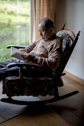 Älterer Mann sitzt im Schaukelstuhl am Fenster und liest ein Buch. - MINF08460