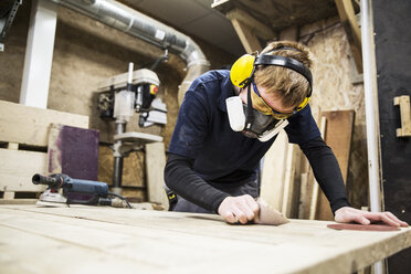 Ein Mann mit Gehörschutz, Schutzbrille und Staubmaske steht in einer Lagerhalle und arbeitet an einem Stück Holz. - MINF08471