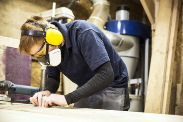 Man wearing ear protectors, protective goggles and dust mask standing in a warehouse, working on a piece of wood. - MINF08472