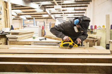 Ein Mann mit Gehörschutz, Schutzbrille und Staubmaske steht in einer Lagerhalle und schleift Bretter aus recyceltem Holz. - MINF08475