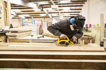 Man wearing ear protectors, protective goggles and dust mask standing in a warehouse, sanding planks of recycled wood. - MINF08475