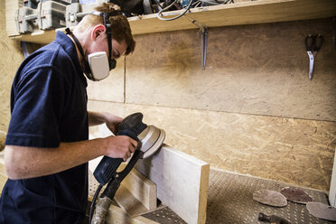 Young man wearing dust mask and protective goggles standing in a workshop, using sander to smoothen edge of piece of wood. - MINF08477