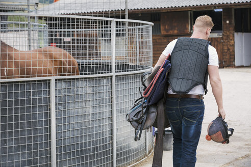 Rear view of a young man wearing a black body protector, and carrying riding gear at a riding stable. - MINF08523