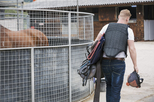 Rückansicht eines jungen Mannes, der einen schwarzen Körperschutz trägt und in einem Reitstall eine Reitausrüstung mit sich führt. - MINF08523