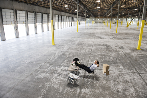 Caucasian man sitting with feet up on desk in front of loading dock doors in a new warehouse interior. stock photo