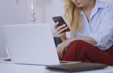 Blonde Frau sitzt auf der Couch und benutzt Laptop und Smartphone - AZF00068