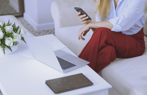 Blonde Frau sitzt auf der Couch und benutzt Laptop und Smartphone, lizenzfreies Stockfoto