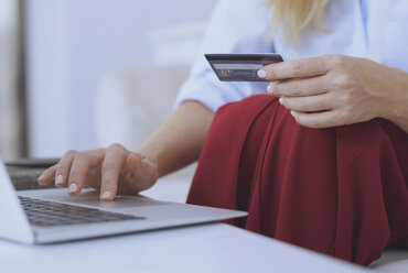 Blonde Frau sitzt auf der Couch und benutzt einen Laptop, um mit ihrer Kreditkarte zu bezahlen - AZF00071