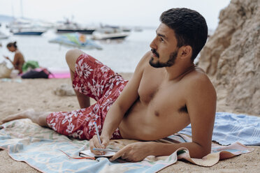 Young man lying on towel at the beach, notebook, thoughtful - AFVF01389