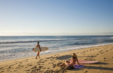 Ein Mann trägt ein Surfbrett, während eine junge Frau im Bikini den Sonnenuntergang über dem Meer genießt. - AURF00258