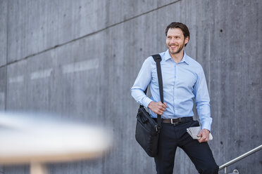 Portrait of smiling businessman with laptop bag at concrete wall - DIGF04805
