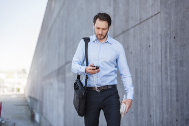 Businessman with earbuds walking along concrete wall looking at smartphone - DIGF04806