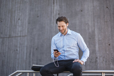 Smiling businessman with earbuds sitting on a railing looking at smartphone - DIGF04808