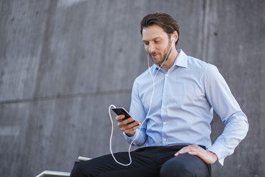 Businessman with earbuds sitting on a railing looking at smartphone - DIGF04809