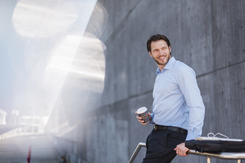 Lächelnder Geschäftsmann mit Laptoptasche und Kaffee zum Mitnehmen lehnt an einem Geländer, lizenzfreies Stockfoto