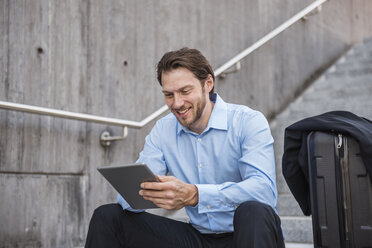 Smiling businessman with rolling suitcase sitting on stairs using tablet - DIGF04817