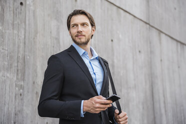 Businessman standing at concrete wall holding smartphone - DIGF04826