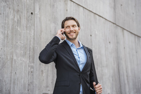 Lächelnder Geschäftsmann an einer Betonwand, der mit seinem Smartphone spricht, lizenzfreies Stockfoto