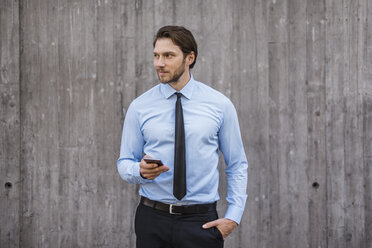 Businessman standing at concrete wall holding smartphone - DIGF04847