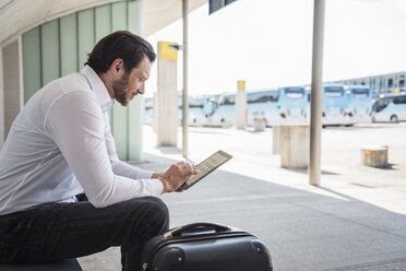Smiling businessman with suitcase sitting at bus terminal using tablet - DIGF04848