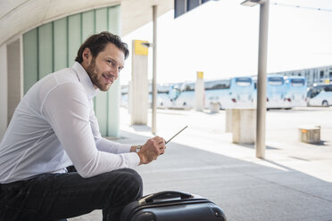 Lächelnder Geschäftsmann mit Koffer sitzt am Busbahnhof und benutzt ein Tablet - DIGF04849