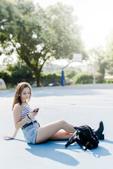 Junge Frau mit Rucksack, die auf einem Sportplatz sitzt und ein Mobiltelefon benutzt - GIOF04146