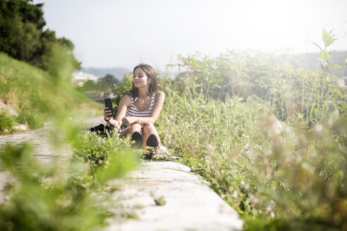 Junge Frau sitzt in der Natur und benutzt ein Mobiltelefon - GIOF04176