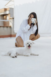Porträt eines weißen Hundes, der auf einer Dachterrasse liegt, während eine junge Frau im Hintergrund ein Foto macht - AFVF01410