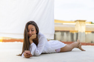 Porträt einer lächelnden jungen Frau, die auf einer Dachterrasse liegt und auf ihr Handy schaut - AFVF01417