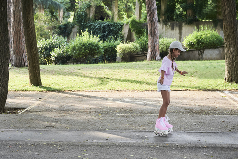 Kleines Mädchen mit Zöpfen und Mütze beim Rollschuhlaufen im Park, lizenzfreies Stockfoto