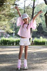 Glückliches kleines Mädchen hebt die Arme beim Rollschuhlaufen im Park - IGGF00514
