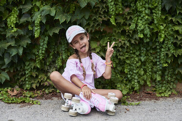 Selbstbewusstes kleines Mädchen, das mit rosa Rollschuhen auf dem Boden sitzt und die Zunge herausstreckt - IGGF00518