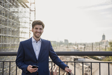 Porträt eines lächelnden Geschäftsmannes, der auf einer Brücke in der Stadt steht und ein Mobiltelefon hält - DIGF04879