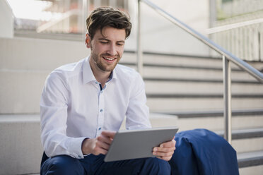 Smiling businessman sitting on stairs in the city using tablet - DIGF04891