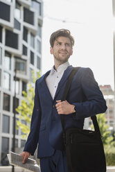 Smiling businessman with laptop bag and newspaper in the city looking around - DIGF04897