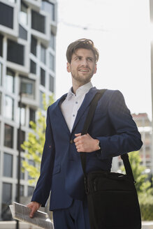 Lächelnder Geschäftsmann mit Laptoptasche und Zeitung in der Stadt, der sich umschaut - DIGF04897