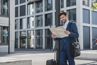 Smiling businessman with baggage reading newspaper in the city - DIGF04898