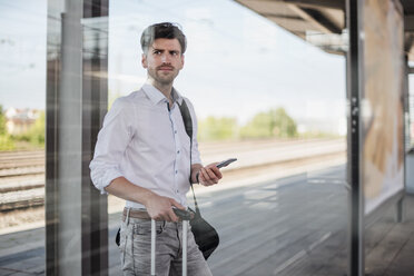 Businessman with baggage and cell phone standing on station platform looking around - DIGF04916