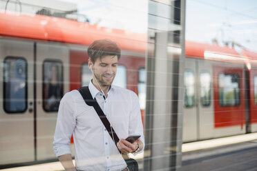 Smiling businessman on station platform with earphones and cell phone - DIGF04920
