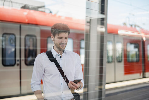 Lächelnder Geschäftsmann auf dem Bahnsteig mit Kopfhörern und Mobiltelefon - DIGF04920
