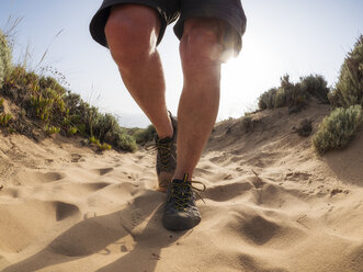 Senior man with hiking shoes walking in sand against the sun - LAF02071