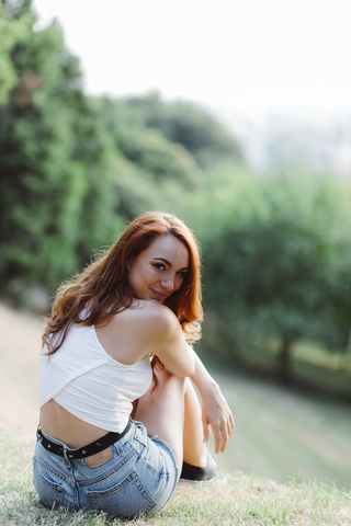 Lächelnde rothaarige Frau entspannt sich auf einem Hügel in einem Park, lizenzfreies Stockfoto