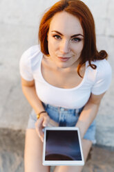 Porträt einer lächelnden rothaarigen Frau, die mit einem digitalen Tablet an der Wand sitzt - GIOF04225
