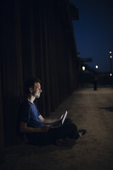 Mid adult man sitting cross-legged on ground, using laptop at night - GUSF00984