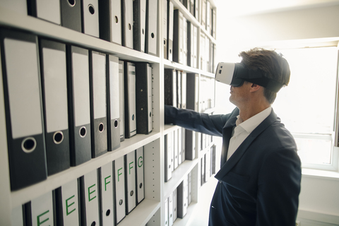 Geschäftsmann mit VR-Brille im Firmenarchiv, sucht Regal mit Akten, lizenzfreies Stockfoto