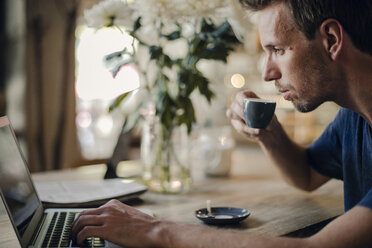 Mid adult man sitting in coffee shop, drinking coffee, using laptop - GUSF01092