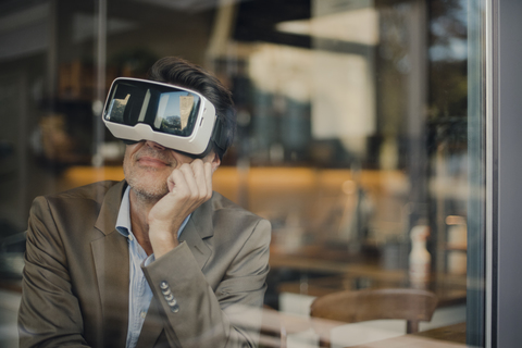 Älterer Geschäftsmann sitzt in einem Café und schaut durch eine VR-Brille, lizenzfreies Stockfoto