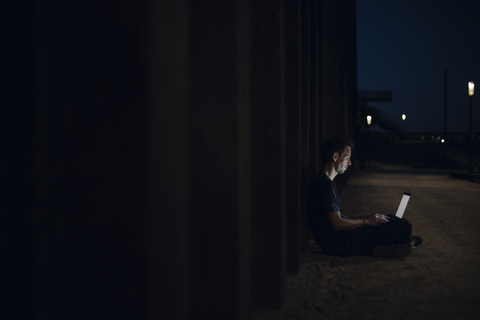 Mittlerer erwachsener Mann im Schneidersitz auf dem Boden sitzend, der nachts einen Laptop benutzt, lizenzfreies Stockfoto