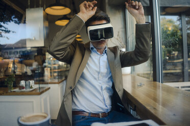 Mature businessman sitting in coffee shop, looking through VR glasses, dancing - GUSF01175