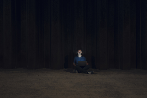 Mid adult man sitting cross-legged on ground, using laptop at night stock photo